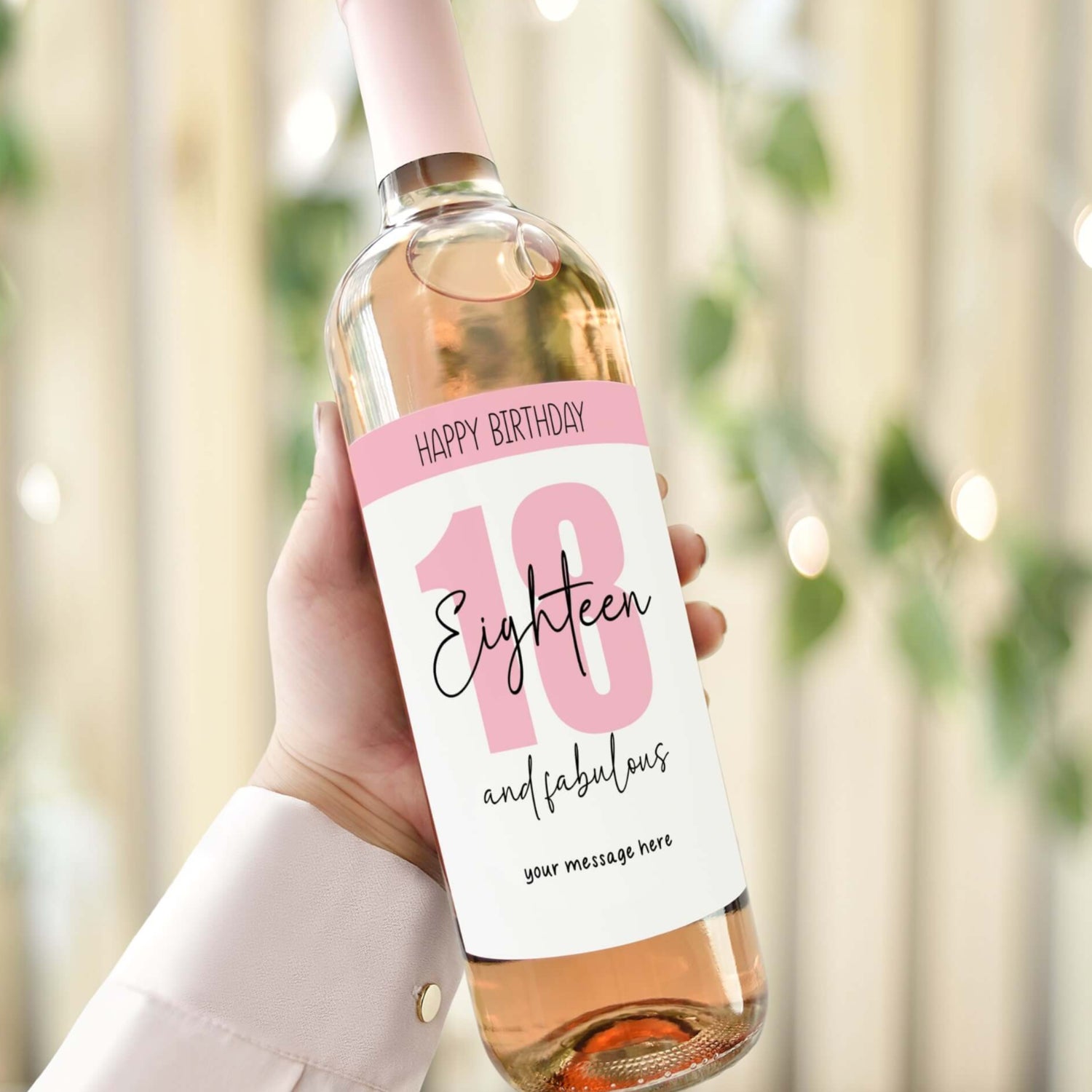 Personalised 18th birthday wine label with 'Eighteen and Fabulous' design in pink and white, held against a party backdrop.