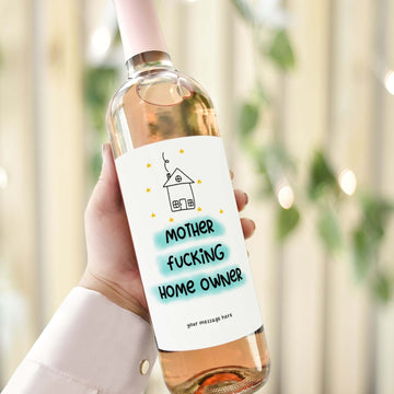 A wine bottle with a funny housewarming label reading "Mother F***ing Home Owner," designed as a gift for celebrating new homeowners.