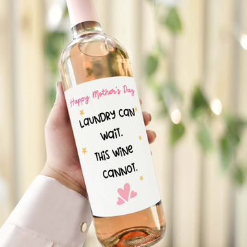 A hand holding a wine bottle with a funny Mother's Day wine label that reads, "Laundry can wait. This wine cannot." Perfect gift for Mum.